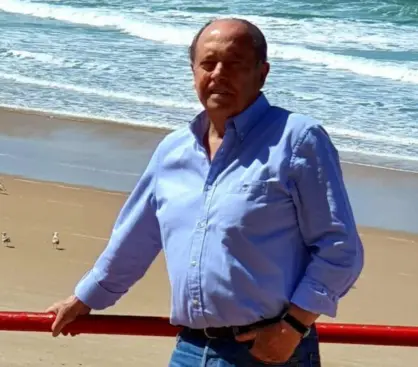 Una fotografía de Jaime Rocha, vestido con una camisa azul y jeans, parado en una playa con el mar de fondo y una barandilla roja en primer plano.